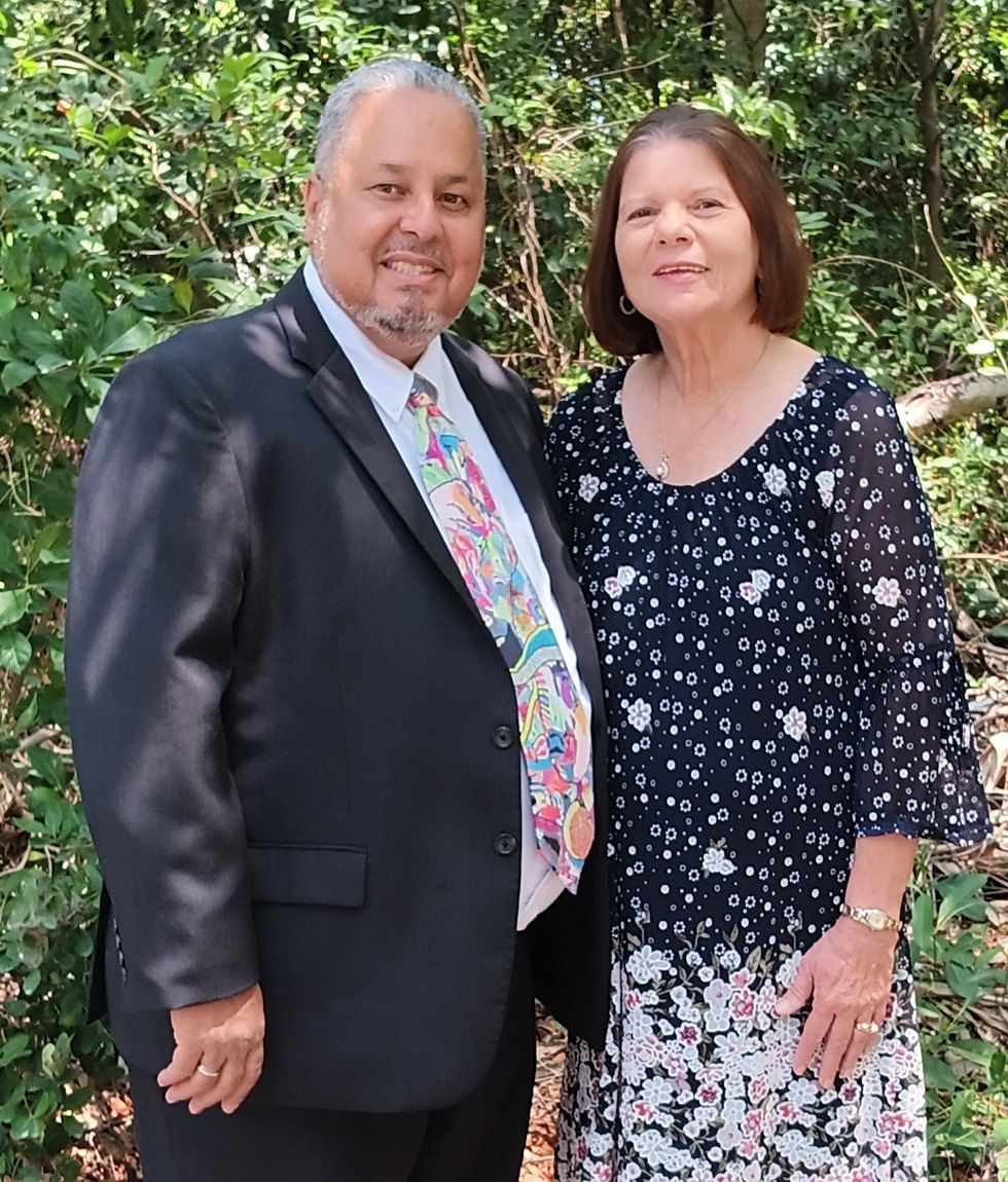 Rick and Donna Fernandez, founders of AFCO 5:20, standing together outdoors in front of dense green foliage.
