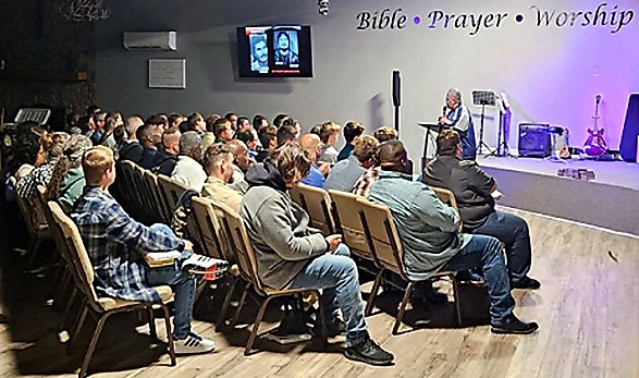 A speaker addresses a room of seated men at a support-group gathering, beneath a wall reading Bible, Prayer, Worship.