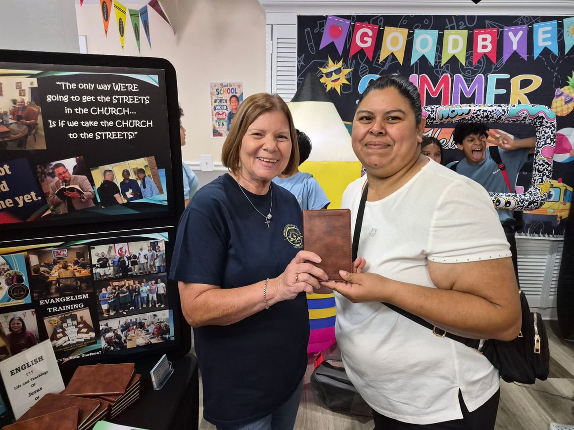 An AFCO 5:20 volunteer and a smiling woman hold a Bible together beside the ministry outreach display at a community event.