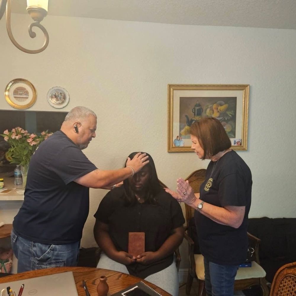 Two AFCO 5:20 volunteers pray over a seated woman during a home visit, one resting a hand on her head while the other prays with open hands.