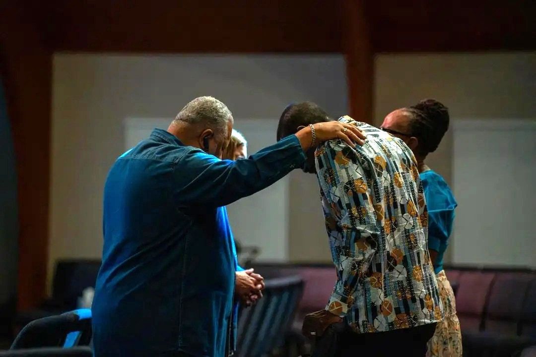 A small group gathers to pray in a church sanctuary in the evening, one man resting a hand on the shoulder of another whose head is bowed.