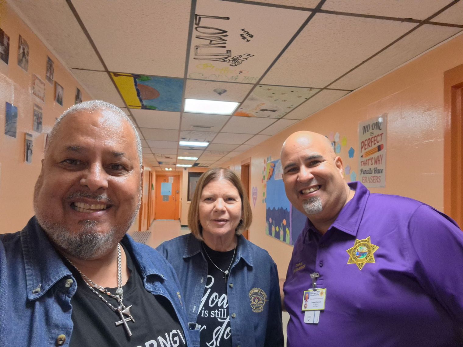 Rick and Donna Fernandez with the chaplain in a facility hallway decorated with artwork and encouraging posters.