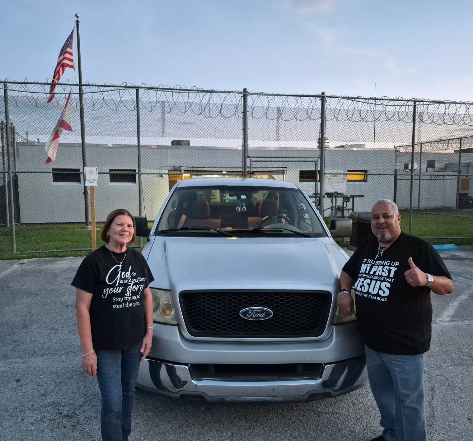 Rick and Donna Fernandez beside their truck outside the facility fence at dusk, an American flag flying overhead.