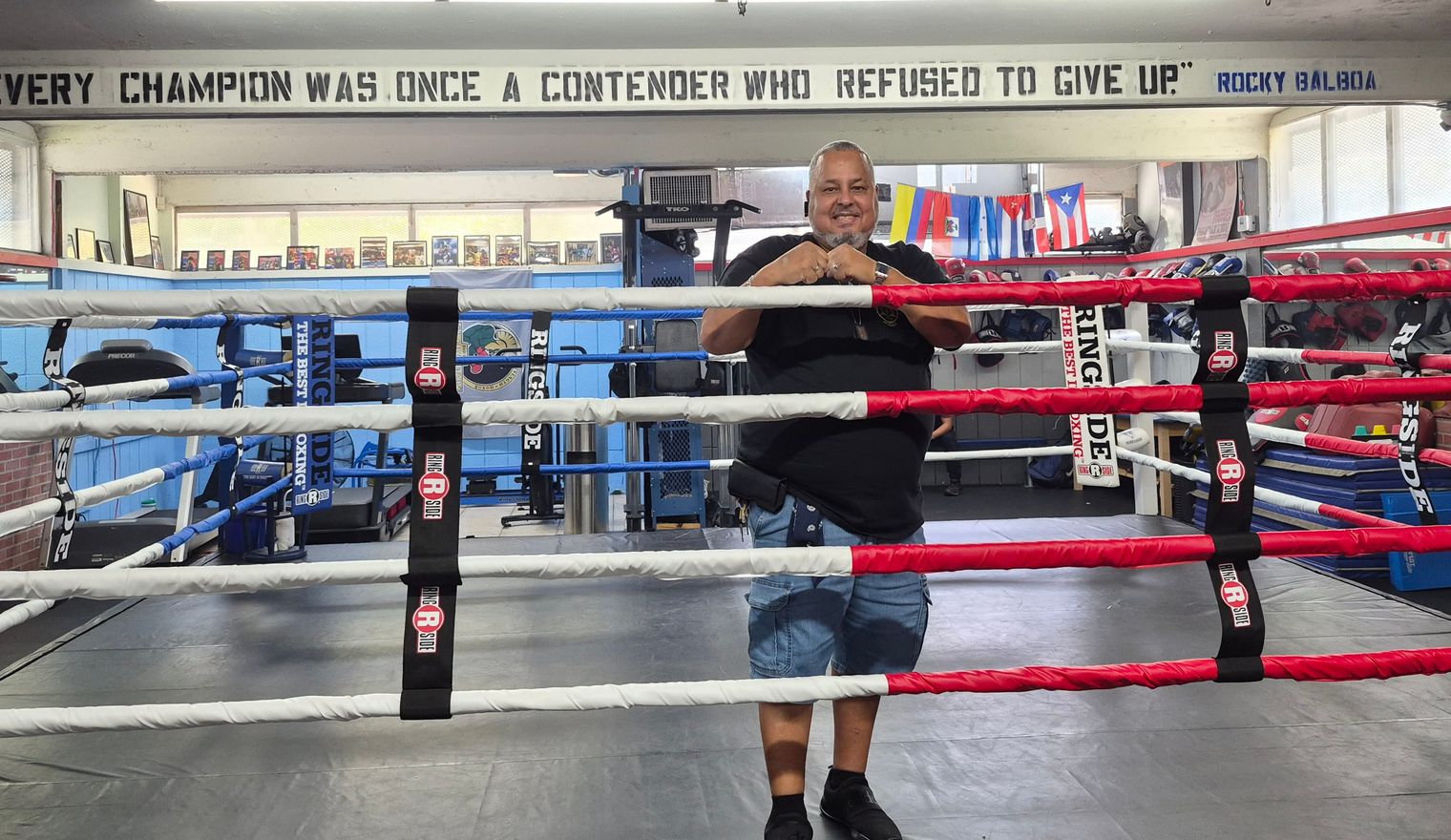 Rick stands in the boxing ring at the PAL gym, beneath a painted quote reading Every champion was once a contender who refused to give up.