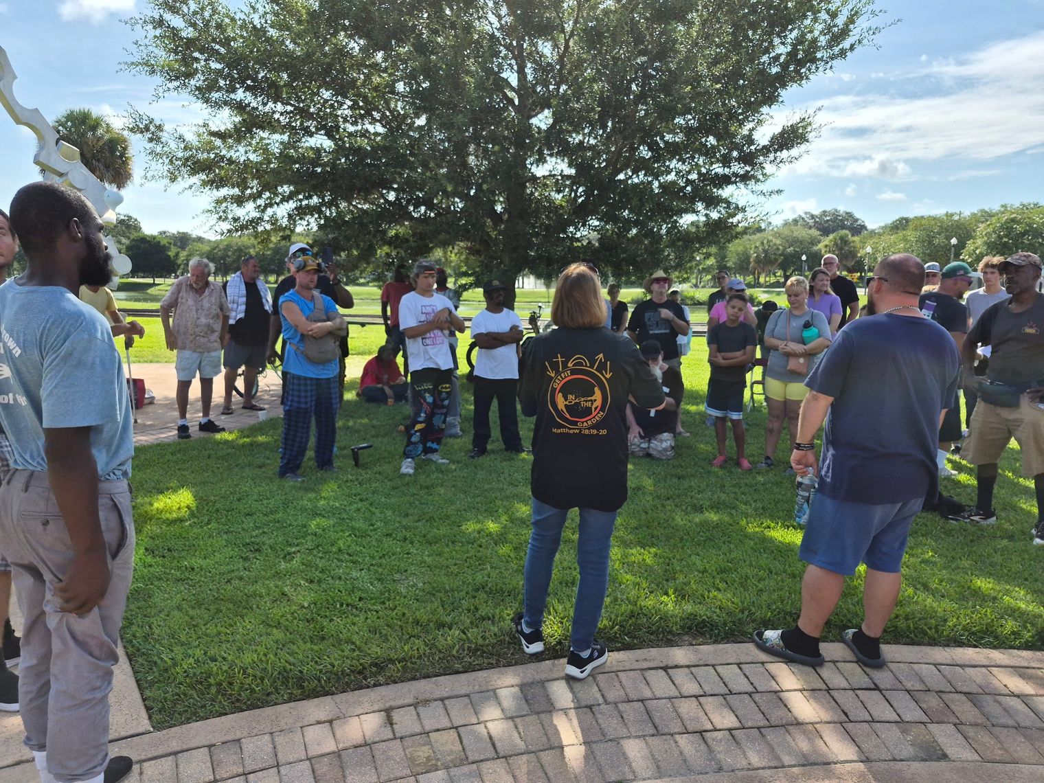 Donna Fernandez, wearing a Get Fit in the Garden shirt, speaks to a crowd gathered under a tree in the park.