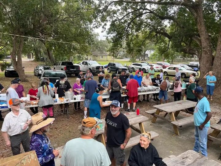 A long serving table under the oaks as neighbors line up for the community meal.
