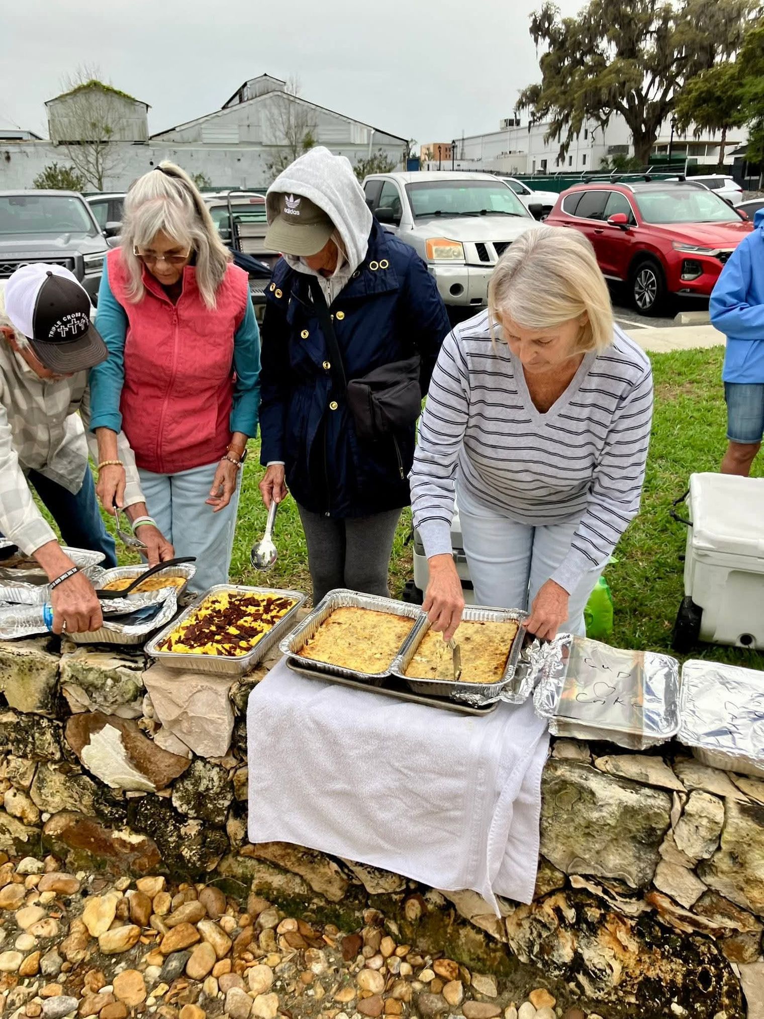 Volunteers serve trays of hot food arranged along a stone wall.
