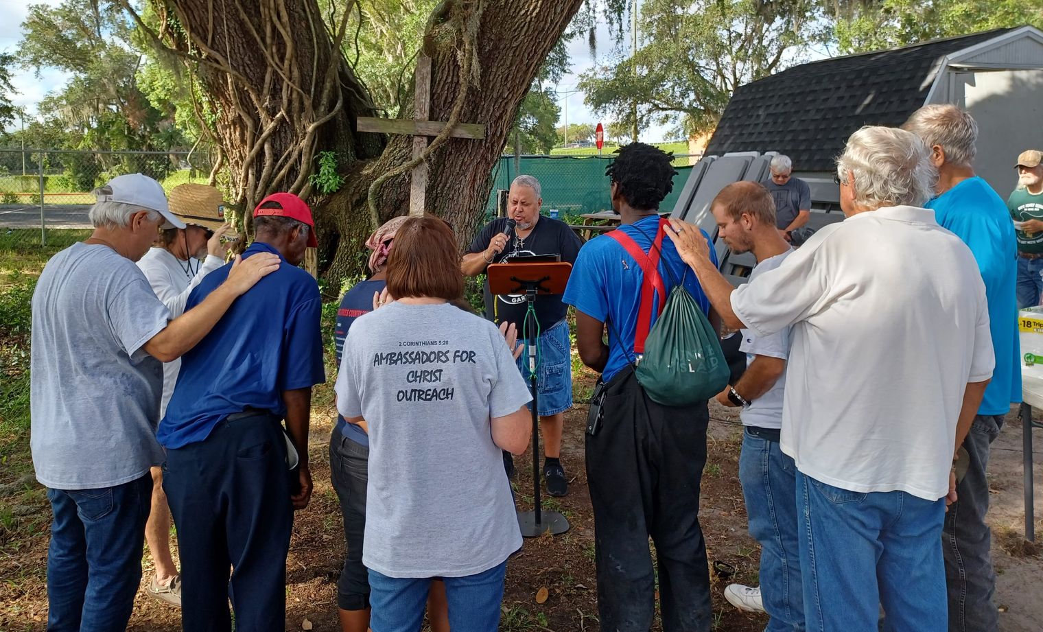 Rick Fernandez prays at the lectern under the oak as the circle lays hands on one another, a volunteer shirt reading Ambassadors for Christ Outreach.